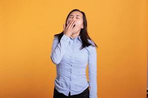 Portrait of sleepy filipino woman covering mouth with hand being tired while posing in studio standing over yellow background. Exhausted model yawning having lazy expression, working overtime photo