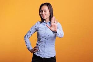 Serious filipino woman doing rejection gesture with hand in front of camera, posing in studio over yellow background. Negative model showing stop gesture, being in denial and expressing refusal. photo