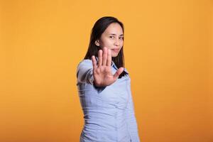 Filipino young adult doing no sign with palm on camera, expressing disapproval and denial. Cheerful woman showing rejection and refusal gesture standing in studio over yellow background photo