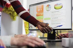 African american employee in festive ornate office showing colleagues graphic financhial charts on computer screen. Business team working together on finishing project tasks before arriving deadline photo