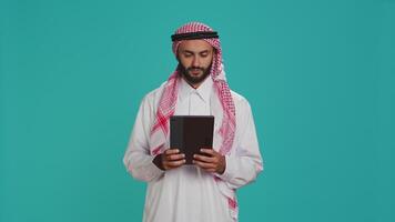 milieu est homme vérification tablette dans studio, navigation sites Internet en ligne sur numérique gadget. Masculin la personne dans arabe vêtements en utilisant dispositif à naviguer sur l'Internet pages, envoyer des SMS sur social médias. video