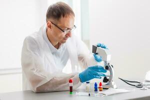 A male doctor or scientist looking through a microscope on a table with laptop computer in background photo