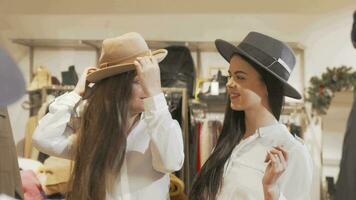 fêmea amigos tendo Diversão às roupas loja tentando chapéus juntos video