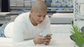 Young African man using smart phone while trying new bed at furniture store video