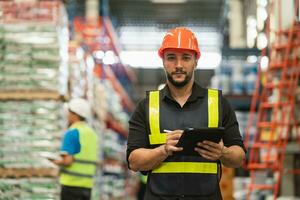 Professional manager man employee smile using tablet check stock working at warehouse. Worker wearing high visibility clothing and a hard hat, helmet and checking and count up goods boxes delivery. photo