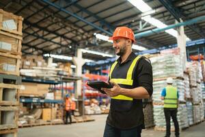 Professional manager man employee smile using tablet check stock working at warehouse. Worker wearing high visibility clothing and a hard hat, helmet and checking and count up goods boxes delivery. photo