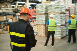 Back side. Professional manager man employee using tablet check stock working at warehouse. Worker ware high visibility clothing and a hard hat, helmet and checking and count up goods boxes delivery. photo