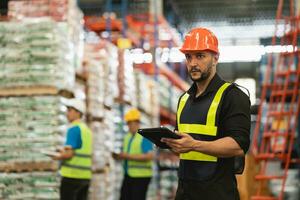 Professional manager man employee using tablet check stock working at warehouse. Worker wearing high visibility clothing and a hard hat, helmet and checking and count up goods or boxes for delivery. photo