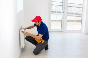 Young handsome professional plumber worker installing heating radiator in an empty room of a newly built apartment or house. Construction, maintenance and repair concept. photo