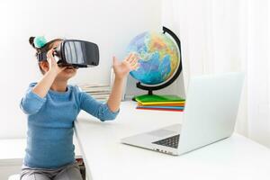 Little kid schoolgirl sit and study at white desk with pc laptop. School distance education at home during quarantine concept. Watching in headset, virtual glasses photo