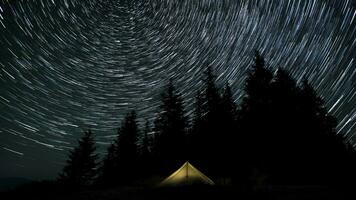 Time lapse of cometshaped star trails in the night sky above a glowing tent and the forest. Epic video 4K