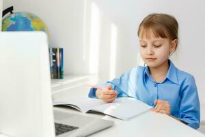 Cute little girl is sitting at table with her laptop and studying online photo