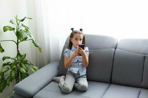 distancia aprendiendo, en línea educación para niños. pequeño niña estudiando a hogar en frente de el teléfono inteligente niño acecho en línea caricaturas, niños computadora adiccion, de los padres control. cuarentena a hogar foto