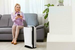 Young woman relaxing on the couch while air humidifier or purifier working on the foreground. Controlling it with a smartphone remotely photo