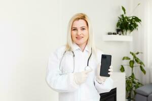 lado perfil retrato sonriente hembra doctor, cuidado de la salud profesional en blanco laboratorio Saco con estetoscopio, analizando datos resultados en móvil inteligente teléfono en pie en hospital pasillo corredor foto