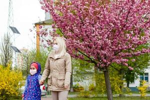 un europeo madre en un respirador con su hija son en pie cerca floración árbol. protector máscara a salvar sí misma desde virus. foto