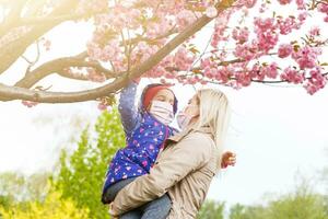 A European mother in a respirator with her daughter are standing near flowering tree. protective mask to save herself from virus. photo