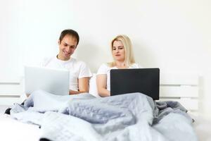 man and woman sitting on bed in morning having fun, laughing, working on laptop onile education, online freelancer job, smiling happy, family living together, bedroom, wearing pajamas photo