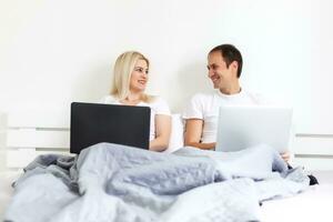 man and woman sitting on bed in morning having fun, laughing, working on laptop onile education, online freelancer job, smiling happy, family living together, bedroom, wearing pajamas photo