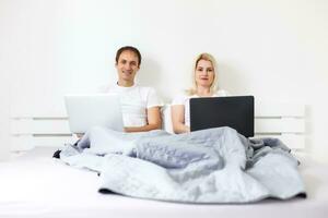 man and woman sitting on bed in morning having fun, laughing, working on laptop onile education, online freelancer job, smiling happy, family living together, bedroom, wearing pajamas photo