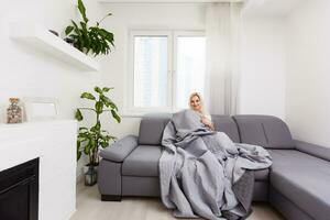 Pretty young woman sitting on sofa covered with blanket, photo