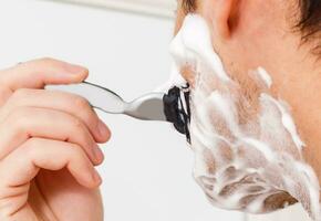 young man shaving in the bath. He is passing the razor for the beard while it looks at the mirror photo