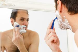 young man shaving in the bath. He is passing the razor for the beard while it looks at the mirror photo