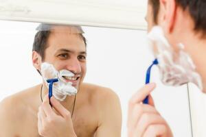 joven hombre afeitado en el baño. él es paso el maquinilla de afeitar para el barba mientras eso mira a el espejo foto