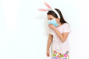 Little caucasian girl with bunny ears and medical mask on her face on a white background. Easter concept. Coronavirus protected photo