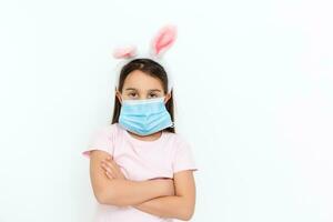 Little caucasian girl with bunny ears and medical mask on her face on a white background. Easter concept. Coronavirus protected photo