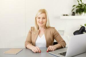 retrato de contento experto medio Envejecido mujer vida entrenador, negocio consultor, psicólogo o médico tutor sonriente alegremente a cámara, trabajando en computadora portátil, disfrutando su trabajo, Ayudar personas en línea foto