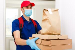 entrega hombre empleado en rojo gorra camiseta uniforme máscara guantes dar comida orden Pizza cajas aislado en amarillo antecedentes estudio. Servicio cuarentena pandemia coronavirus virus gripe 2019-ncov concepto foto