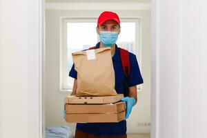 entrega hombre empleado en rojo gorra camiseta uniforme máscara guantes dar comida orden Pizza cajas aislado en amarillo antecedentes estudio. Servicio cuarentena pandemia coronavirus virus gripe 2019-ncov concepto foto