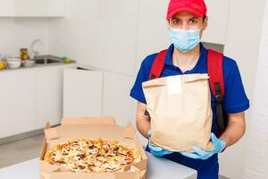 entrega hombre empleado en rojo gorra camiseta uniforme máscara guantes dar comida orden Pizza cajas aislado en amarillo antecedentes estudio. Servicio cuarentena pandemia coronavirus virus gripe 2019-ncov concepto foto