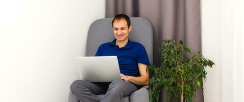 contento joven hombre en camiseta sentado en sofá a hogar, trabajando en ordenador portátil computadora, sonriente. foto