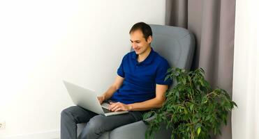 contento joven hombre en camiseta sentado en sofá a hogar, trabajando en ordenador portátil computadora, sonriente. foto