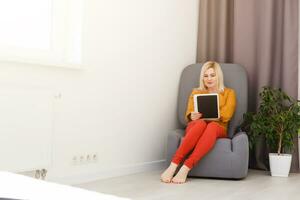 mid adult woman sitting on sofa with tablet pc. Horizontal shape, front view, copy space photo