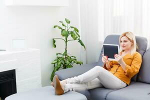 mid adult woman sitting on sofa with tablet pc. Horizontal shape, front view, copy space photo