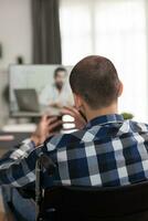 Doctor instructions during video call for disabled man in wheelchair. Young immobilized guy sitting at home in front of laptop, talking with his doctor about the process of rehabilitation on video call photo