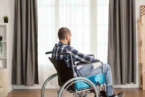 Young depressed sitting in wheelchair near window at home. Serious sad caucasian man wearing casual clothes and look at large panoramic view in bright modern living room, hospital or clinic. photo