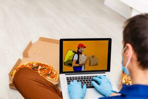 hombre en protector guantes y máscara utilizando ordenador portátil para en línea comida orden durante cuarentena, de cerca. entrega Servicio foto