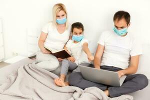 Family in medical face masks lying on a bed with a laptop. Concept of watching video, working together and quarantine during a COVID-19 coronavirus epidemic photo