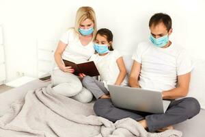 Family in medical face masks lying on a bed with a laptop. Concept of watching video, working together and quarantine during a COVID-19 coronavirus epidemic photo