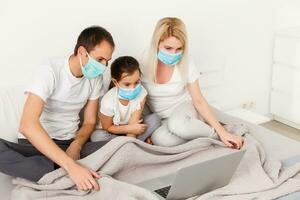 Family in medical face masks lying on a bed with a laptop. Concept of watching video, working together and quarantine during a COVID-19 coronavirus epidemic photo