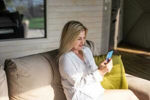 Happy young caucasian woman relax on comfortable couch at home texting messaging on smartphone, use cellphone, browse wireless internet on gadget, shopping online from home photo