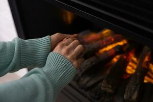 mujer frotamiento manos y calefacción en frente un fuego sitio a hogar en invierno. foto