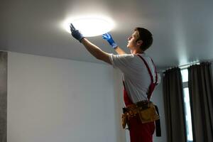 Electrician at work. Service for the repair of electrical wiring and replacement of ceiling lamps. A builder is installing a loftstyle wooden ceiling. Rentagent helps with the housework photo