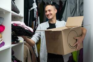 Happy cheerful ethnic male in casual clothes carrying cardboard box and looking at camera on moving day with cartons and drawing easels on blurred background. photo