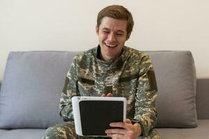 Smiling millennial man in camouflage uniform holding modern digital tablet, chatting with family, posing on white studio background, copy space. Modern technologies and military personnel concept. photo