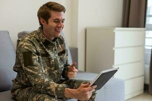 Smiling millennial man in camouflage uniform holding modern digital tablet, chatting with family, posing on white studio background, copy space. Modern technologies and military personnel concept. photo